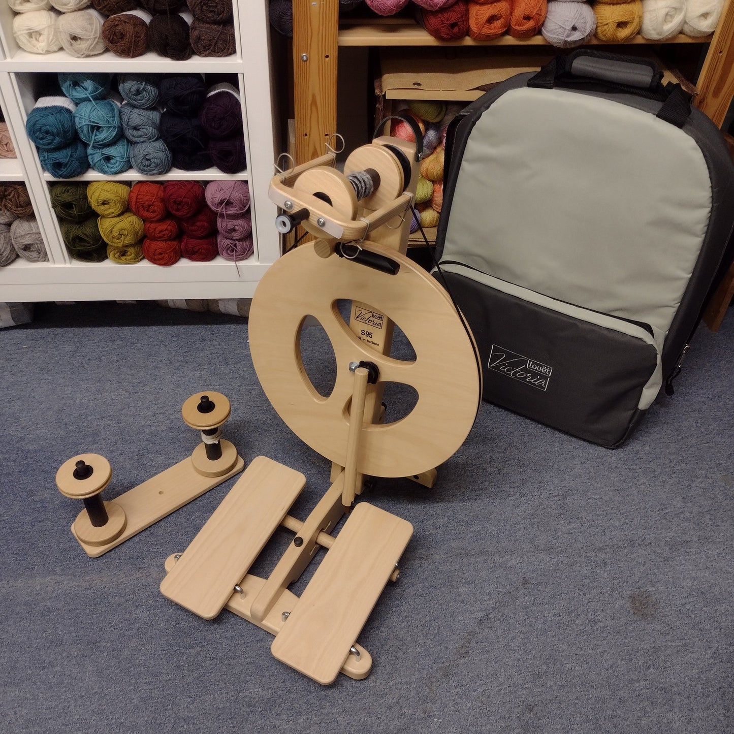 A wooden spinning wheel with parts spread out on a table, alongside a black carrying case, in a workshop setting with yarn and fabric in the background.