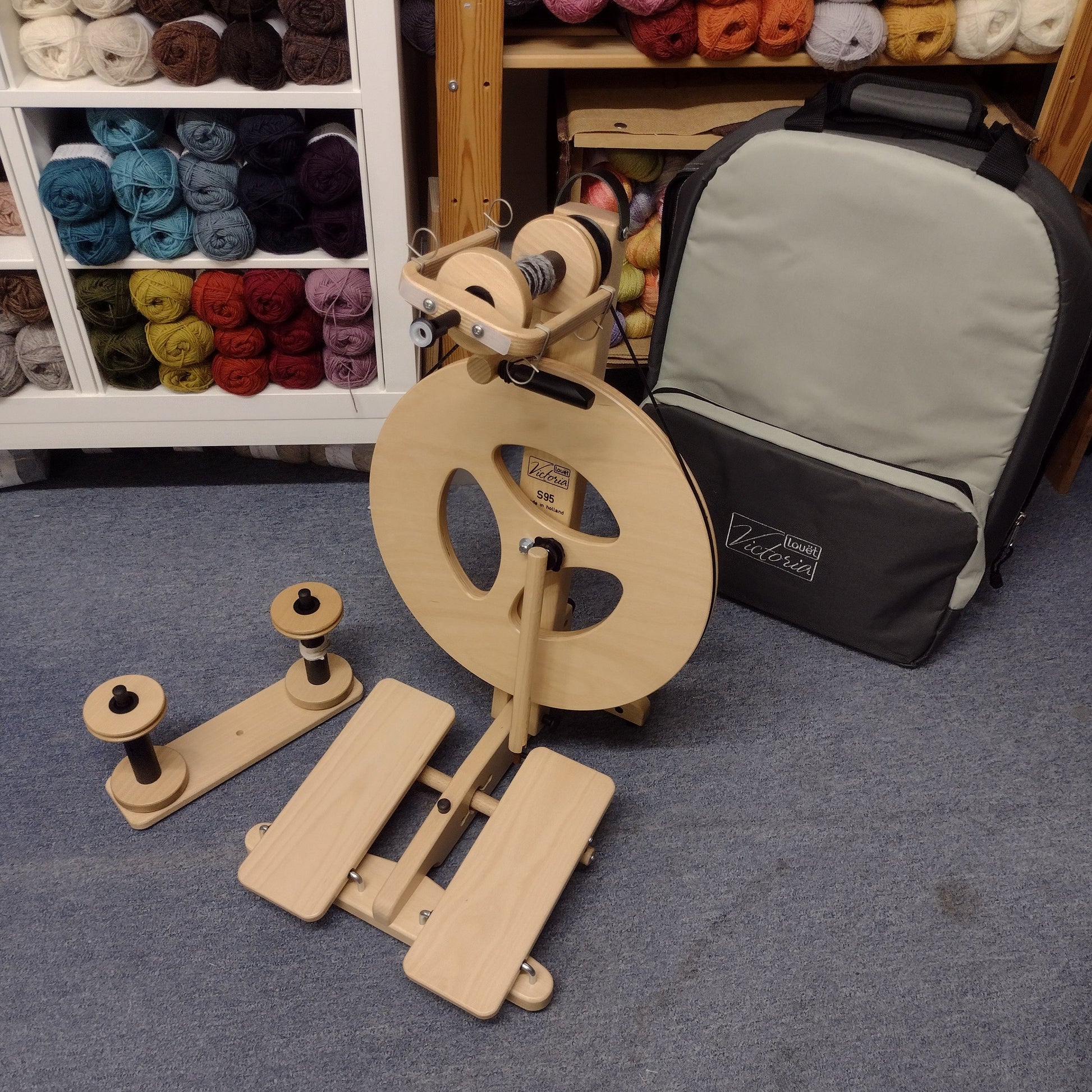 A wooden spinning wheel with parts spread out on a table, alongside a black carrying case, in a workshop setting with yarn and fabric in the background.