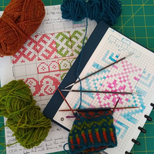 A variety of colorful yarns and knitting needles next to design paper and a notebook with fair isle pattern designs.